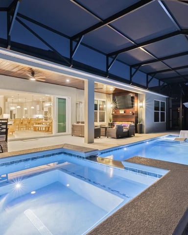 Lit screened pool and spa at night with outdoor seating and open kitchen view