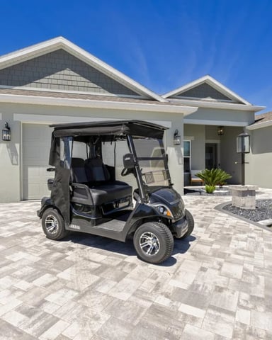 Grey house with black Yamaha golf cart parked on paver driveway with palm trees and blue sky