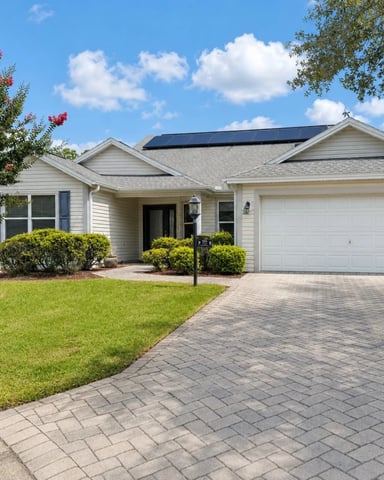 Single-story white house with solar panels, two-car garage, brick driveway, and manicured landscaping with flowering shrubs.