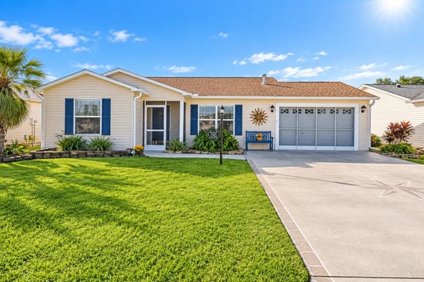 Single-story beige house with blue shutters, two-car garage, and manicured lawn under clear blue sky