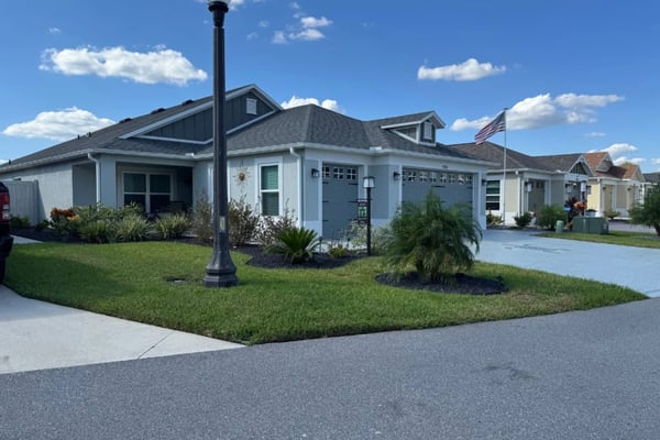 Light blue single-story house with gray roof, two-car garage, and American flag in front yard with palm trees and landscaping.