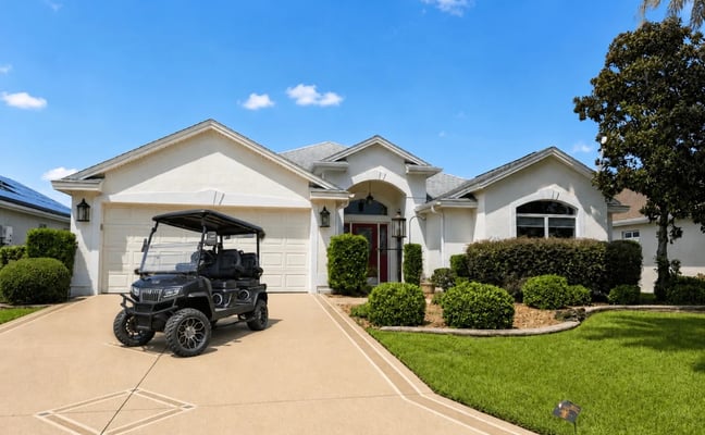 White single-story home with golf cart on driveway, manicured lawn, and landscaped shrubs under clear blue sky